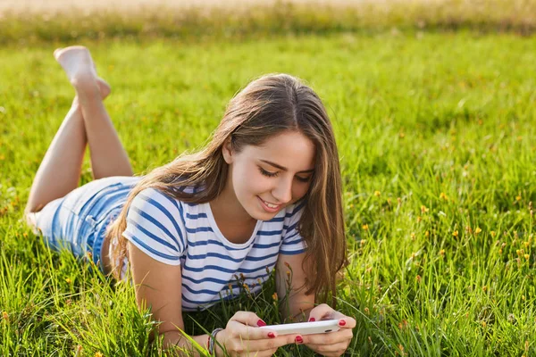 A portrait of young girl with dark long hair wearing striped T-shirt and jeans shorts relaxing on the grass with her legs raised up looking in her smartphone and enjoying nice sunny summer weather — Stock Photo, Image
