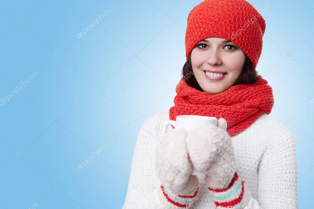 A pretty woman with bright smile and nice make-up wearing knitted warm hat, scarf, sweater and mittens holding a cup of hot delicious tea in her hands. Laughing girl drinking hot tea to warm up