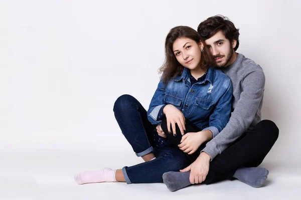 Um casal feliz sentado cruzou as pernas no chão. Um tipo barbudo a abraçar a namorada com amor. Duas pessoas sentadas perto umas das outras descalças isoladas sobre fundo branco — Fotografia de Stock