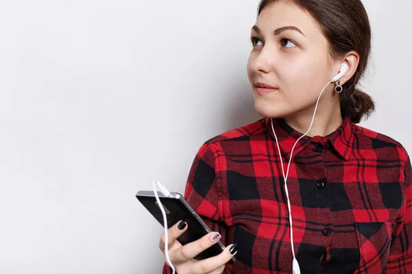 Hipster flicka i röd rutig skjorta att ha håret flätat i en svans som håller mobiltelefon lyssnar på musik eller ljudbok med hörlurar, poserar mot vita studio vägg bakgrund med kopia utrymme — Stockfoto
