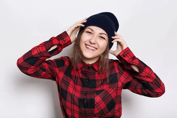 Conceito de juventude e felicidade. Menina adolescente bonita vestindo boné elegante e vermelho verificado camisa sendo feliz e sorrindo segurando as mãos na cabeça.Retrato de feliz atraente elegante jovem fêmea — Fotografia de Stock