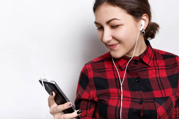 Дівчина-підліток у стильній перевіреній сорочці, яка слухає свою улюблену музику з навушниками, посміхаючись, дивлячись на екран. Красива школярка в навушниках стоїть на білому тлі — стокове фото