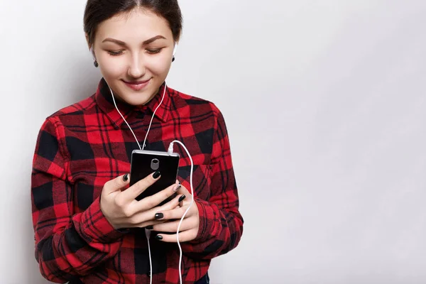 Happy hipster girl mengenakan baju merah diperiksa memegang smartphone di tangannya mendengarkan musik atau buku audio dengan headphone, menerima pesan dari pacarnya tampak bahagia ke layar — Stok Foto