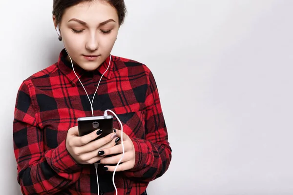 Plan intérieur d'une jolie jeune fille hipster vêtue d'une chemise à carreaux décontractée écoutant un livre audio ou une radio sur un téléphone portable avec écouteurs. . — Photo