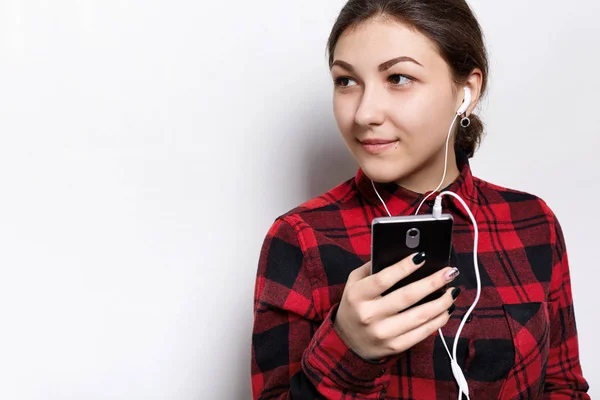 Wanita muda menarik dengan rambut gelap dan kulit berbintik-bintik sehat berpaling dengan ekspresi mempesona, mendengarkan musik favoritnya dengan earphone, berdiri melawan dinding ruang fotokopi putih — Stok Foto