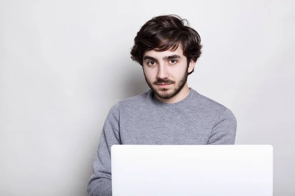 Szép szakállas férfi sylish sötét hajat visel a hétköznapi szürke pulóver, laptop, e-mailek ellenőrzése komoly kifejezés közvetlenül a kamerába nézett, ingyenes vezeték nélküli internet-kapcsolat segítségével — Stock Fotó