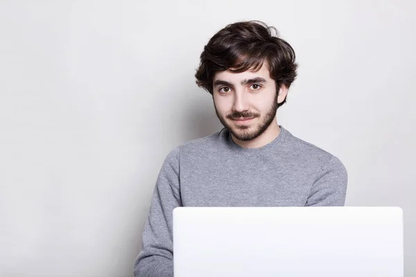 Egy magabiztos vonzó srác sötét szakáll és elegáns frizura előtt nyitott laptop számítógép amelynek bájos mosollyal vizsgálja a kamera, a nagy barna szemek, elszigetelt fehér falon ülve — Stock Fotó