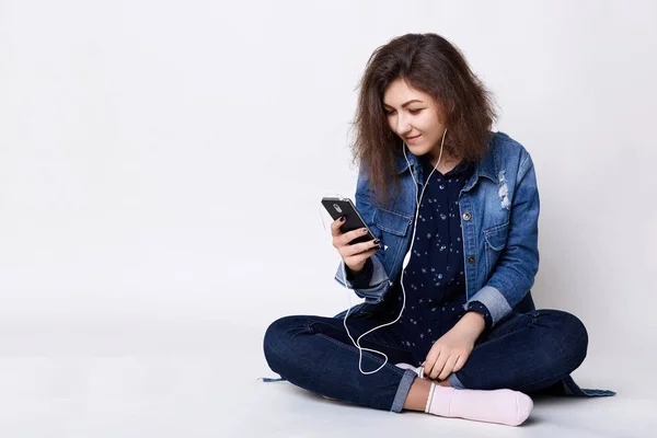 Sebuah gambar gadis menarik mengenakan jaket jean dan jeans duduk santai di lantai kaki menyilang memiliki earphone di telinganya senang untuk menerima pesan dari pacarnya . — Stok Foto