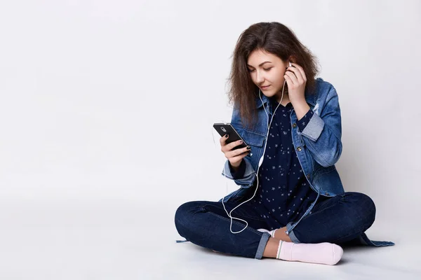 Personas, tecnología y comunicación. Una chica morena bastante joven escuchando la música con su celular sosteniendo su mano en sus auriculares sentados piernas cruzadas en un estudio blanco — Foto de Stock