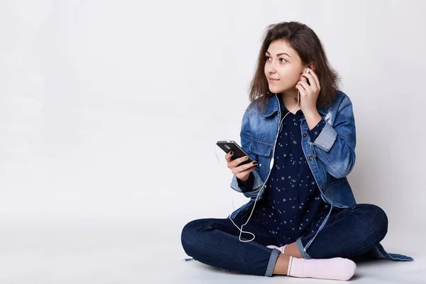 Orang, gaya hidup dan teknologi. Fashionable wanita muda duduk menyilangkan kaki di lantai putih memegang smartphone di tangannya mendengarkan musik mengetik pesan tampak samping senang — Stok Foto