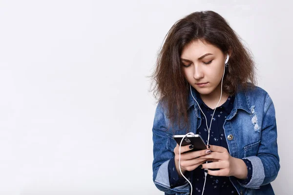 Seorang gadis cantik bergaya muda dengan rambut gelap mengenakan pakaian modern memegang smartphone di tangannya memiliki earphone di telinganya mendengarkan musik yang mencari sesuatu di telepon . — Stok Foto