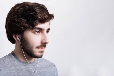 İnsanlar ve yaşam tarzı kavramı. Yana doğru mesafe içine bakarak bazı elektronik cihaz kullanarak beyaz kulaklık müzik dinlerken gri kazak giyen yakışıklı genç sakallı adam portresi 