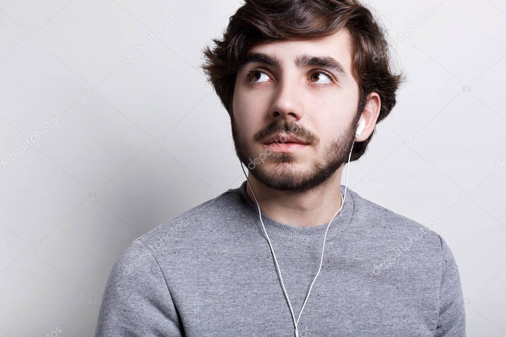 Closeup portrait of handsome bearded man in headphones listening to music via mobile phone dressed in grey casual sweater looking up with thoughtful expression isolated over white background