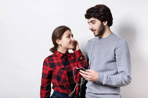 Uma mulher vestida de vermelho verificado camisa e barbudo cara em camisola cinza segurando um smartphone ouvindo a música com fones de ouvido juntos olhando um para o outro com amor. Tecnologia e pessoas . — Fotografia de Stock