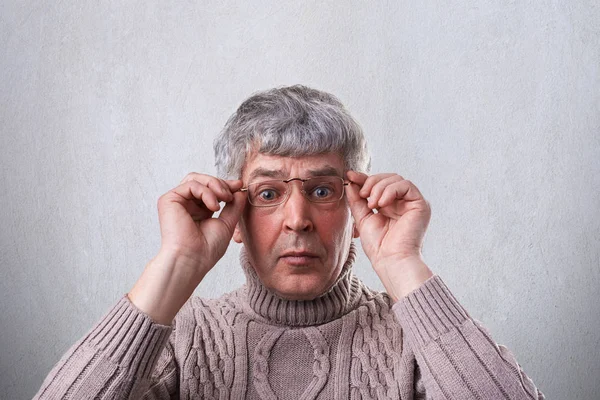 Sebuah close-up dari heran pria senior mengenakan kacamata dan sweater memegang tangannya pada frame kacamata mencari dengan mata terbuka lebar ke kamera. Seorang pria tua yang terkejut atas latar belakang putih — Stok Foto