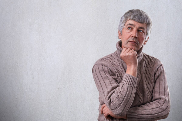 A portrait of handsome senior man standing near white wall dreaming about something looking aside holding his hand under his chin. Pensive grandfather thinking about his life and children. 