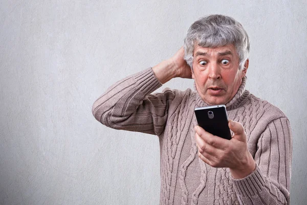 Портрет здивованого старшого чоловіка, який дивиться з широко відкритими очима в свій смартфон, вражає те, що він бачить на своєму мобільному телефоні. Людські вирази обличчя. Сюрприз, дивовижність, диво — стокове фото