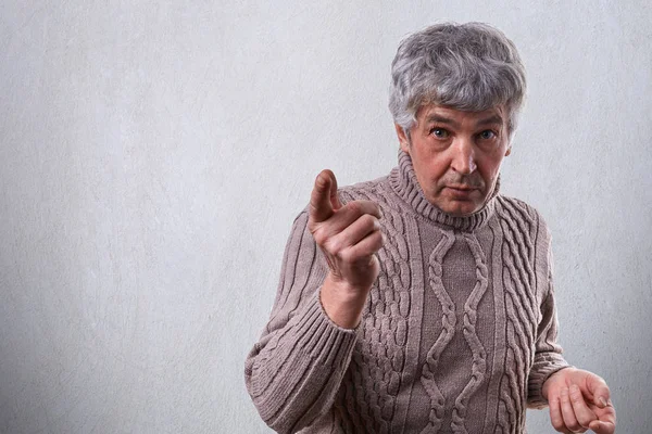 Um homem idoso bonito espantado com cabelo grisalho vestido com suéter em pé perto da parede branca apontando com o dedo querendo dizer algo importante. O senhor ameaça com o dedo. Homem descontente — Fotografia de Stock