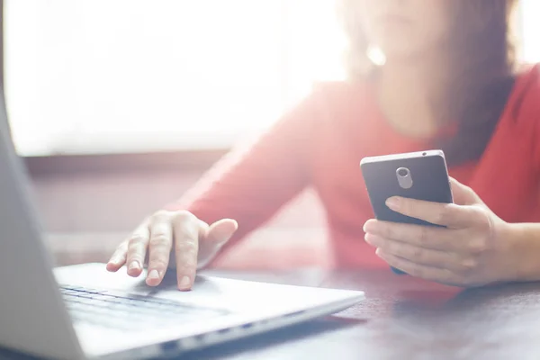 Closeup image of woman`s hands holding smartphone and typing something at keyboard of laptop. Young female searching information in network on mobile phone and laptop during free time. — Stock Photo, Image