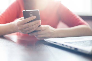 Cep telefonu tutan ve açık dizüstü bilgisayar ile tahta masada otururken mesajlar yazarak genç kadın. Genç kadın alışveriş dizüstü bilgisayar ve akıllı telefon tarafından çevrimiçi yapma. Modern teknoloji ve insanlar.