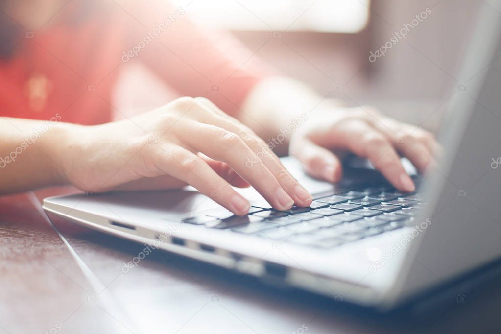 Female hands typing on keyboard of laptop surfing Internet and texting friends via social networks, sitting at wooden table indoors. Tecnology and communication concept/ Selective focus.