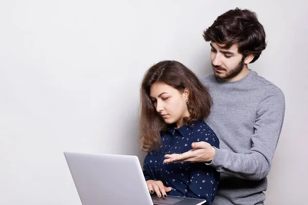 Боковой портрет двух молодых людей, использующих ноутбук. Красивая девушка держит в руках ноутбук для онлайн-покупок. Молодой хипстер стоит позади девушки, помогая ей выбрать, что купить — стоковое фото