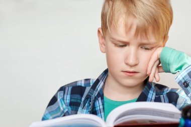 Kitap, okul, çocuk. Sarı saçlı küçük okul çocuğu siting ve ifade yorgun kitap haline bakarak kontrol forması giymiş. Küçük bir çocuk bir kitap okuma dersleri için hazırlanıyor