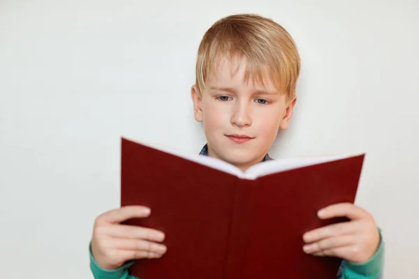 Un ragazzino carino con i capelli biondi e gli occhi azzurri che reggono il rosso ha aperto il libro leggendo storie interessanti. Un bambino di sette anni che legge un libro con grande interesse. Concetto di persone e lifestyle . — Foto Stock