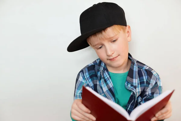 A portrait of curious little boy in black cap and checked colorful shirt reading encyclopedia looking into the book with interest. Education,  school, learning and people concept. — Stock Photo, Image