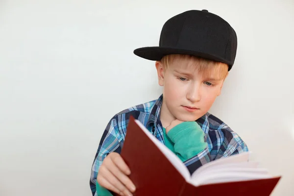小学年龄做家庭作业的孩子。在帽，金色的头发，蓝眼睛的男孩做功课拿着一本书阅读新材料。回到学校。教育理念. — 图库照片