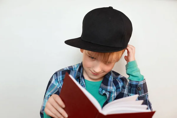 小好奇的孩子与头发的时尚帽挠挠脑袋试图理解什么写在他手里拿着大红色的书一幅画像。儿童和教育. — 图库照片
