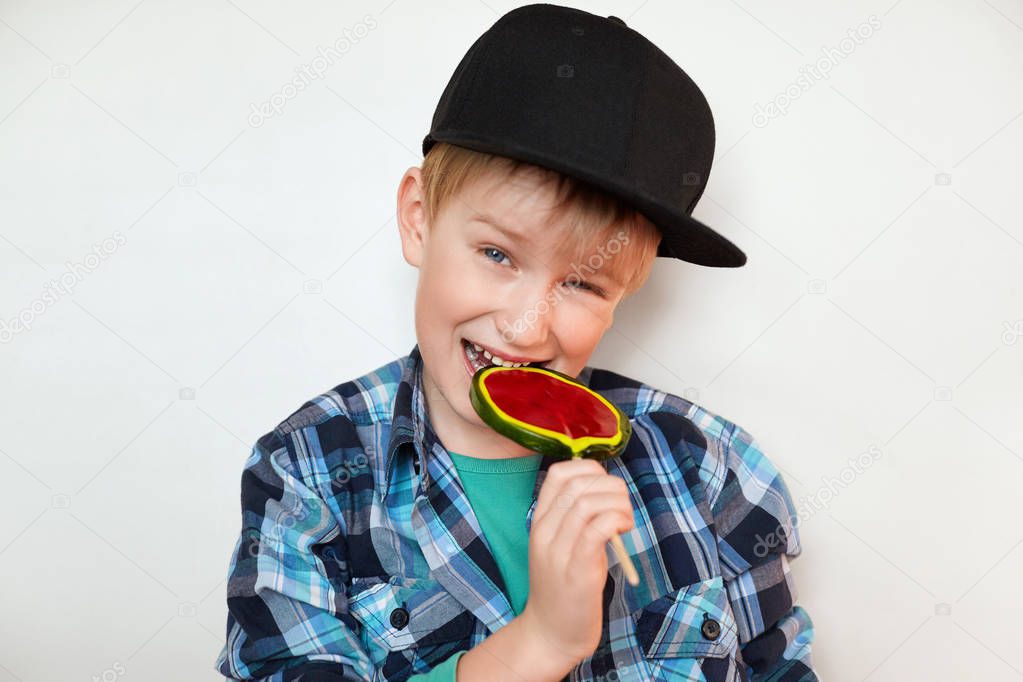 Headshot of beautiful little boy holding and biting lollipop looking at the camera with happy and joyful expression. Handsome boy in stylish clothes isolated over white background with sweet candy 