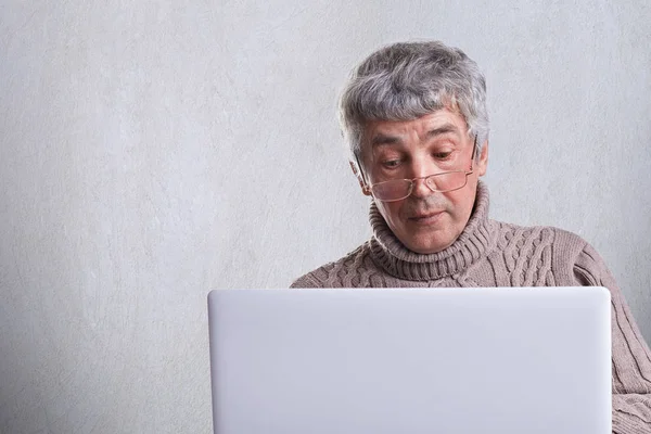 Здивований старший чоловік, одягнений в светр, має сіре волосся і зморшки, використовуючи свій ноутбук серфінг в Інтернеті. Самотній красивий дідусь читає новини онлайн вдома . — стокове фото