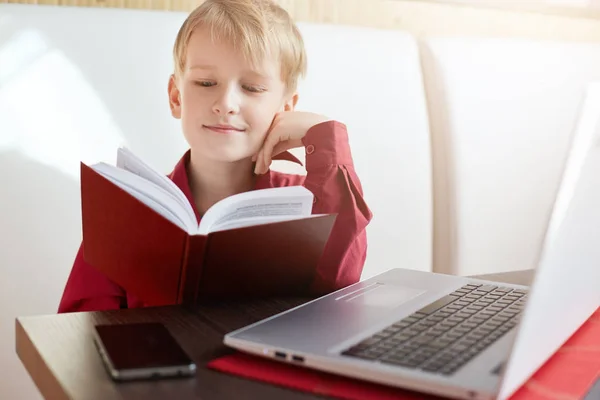 Enfance et loisirs. Portrait d'un petit garçon sérieux assis devant le carnet ouvert lisant un livre avec grand intérêt. Un bel enfant se préparant pour les leçons. Technologie et études . — Photo