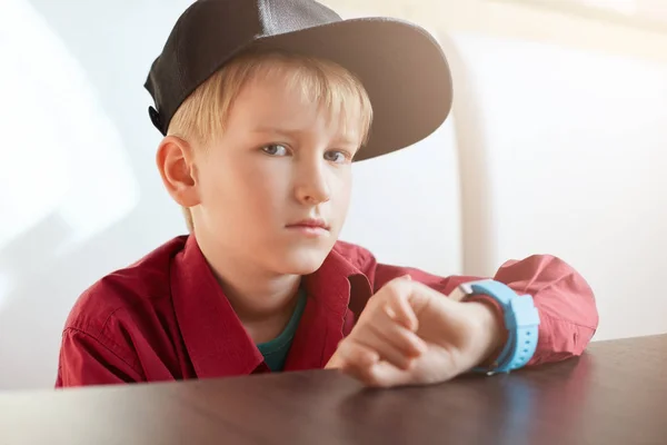 严重的男性儿童穿着时髦的帽子和红色衬衫手腕坐在木桌旁，在白色背景上有一块智能手表地壳画像。一个英俊的小男孩手表. — 图库照片