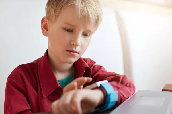 A cute boy with blond hair wearing red stylish shirt sitting on the white couch working with laptop looking at his smart watch touching the screen. A child using electronic device. Lifestyle concept. Stock Photo