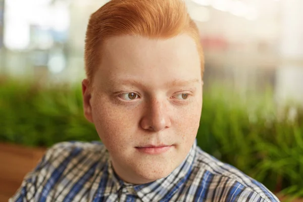 Retrato de cerca de un niño con la cara redonda y muchas pecas con los ojos marrones y el pelo rojo con camisa a cuadros sentado sobre la plantación verde en el café mirando a un lado con expresión pensativa —  Fotos de Stock