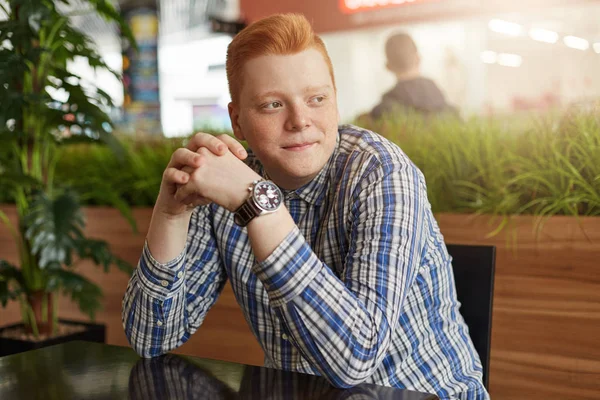 A portrait of redhead man with freckles wearing stylish checked shirt and watch sitting in cosy cafe waiting for his friends to come looking happily aside. Redhead hipster with trendy haircut.