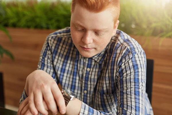 Joven y elegante hombre de cabeza roja vestido con camisa a cuadros que se encuentra en un acogedor café mirando su reloj en espera de alguien. Estilo y concepto de moda. Un joven empresario a la espera del encuentro en la cafetería . — Foto de Stock