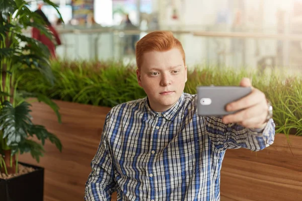 Young freckled redhead man in checked shirt making selfie using mobile phone posing against green plants having serious expression. A stylish man taking photograph with mobile phone looking at camera.
