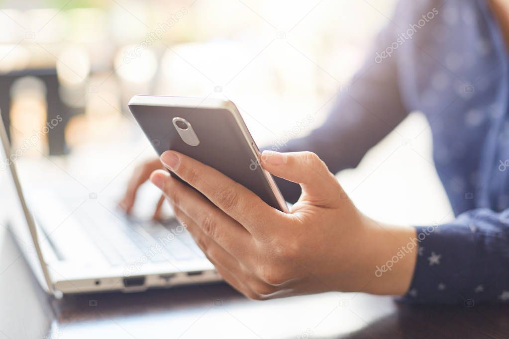 Modern technology and lifestyle concept. A close-up of woman`s hands holding smartphone and typing something at her laptop. A woman having dinner break at cafe using free internet connection. 