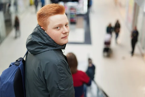 A redhead freckled stylish boy standing back in shopping mall wearing fashionable jacket with hood and holding rucksack on his back doing shopping. People, fashion, lifestyle concept.