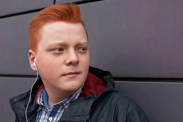 A close-up freckled redhead boy wearing in black jacket and checked shirt having earphones in his ears listening to the music while posing over black wall looking aside