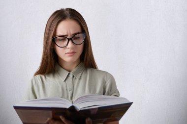 İnsanlar ve eğitim kavramı. Uzun olması ciddi bir genç kız öğrenci kitap siyah saçlı giyen gömlek ve büyük holding gözlük okuma dersleri için hazırlanıyor. Zeki kız bir kitap okuma