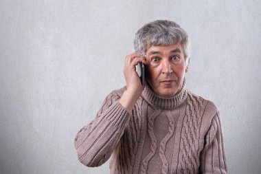 Gri saç olması kazaklı şaşırmış bir yaşlı adam yuvarlak koyu renk gözleri ve kırışıklıklar yüzünde beyaz arka plan karşı dururken telefon üzerinden iletişim. İnsan ve teknoloji kavramı.