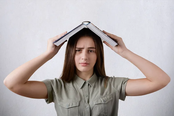 Eine junge Studentin im Hemd mit braunen langen glatten Haaren und dunklen Augen, die ein großes Buch auf dem Kopf hält und nach der Lektüre müde wirkt. Menschen, Bildung, Emotionskonzept. — Stockfoto