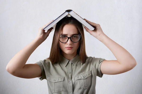 Retrato horizontal de una joven estudiante con anteojos y camisa con libro en la cabeza cansada de estudiar. Hermosa chica trabajando en su proyecto de diploma . —  Fotos de Stock