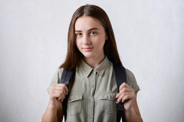 A pretty schoolgirl with long straight hair and dark beautiful eyes wearing elegant shirt holding rucksack on her back isolated over white background going to school. School concept. — Stock Photo, Image
