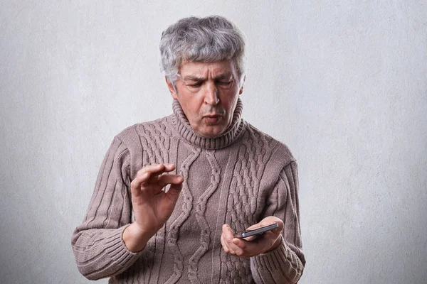 Здивований зрілий чоловік у коричневому светрі тримає смартфон, намагаючись зрозуміти, як його вмикати. Проблемний літній чоловік тримає смартфон, не знаючи, як ним користуватися . — стокове фото