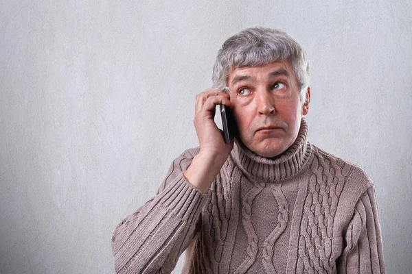 A horizontal portrait of mature man with gray hair and wrinkles dressed in warm sweater holding mobile phone on his ear looking aside while speaking isolated over white background. — Stock Photo, Image
